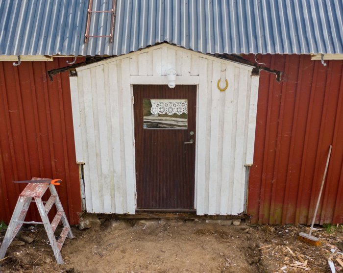 Farstukvist med vitmålade vertikala paneler och brunt dörr, inklämd mellan röda väggar under ett plåttak.