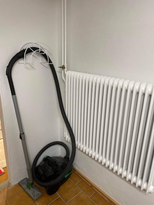 Vit klädhängare på vägg bredvid en radiator med dammsugare och munstycke lutade mot hängaren.