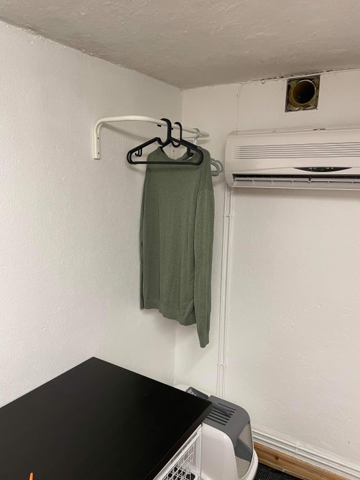 Vit klädstång på vit vägg med svarta klädhängare och grön tröja, intill luftkonditionering.