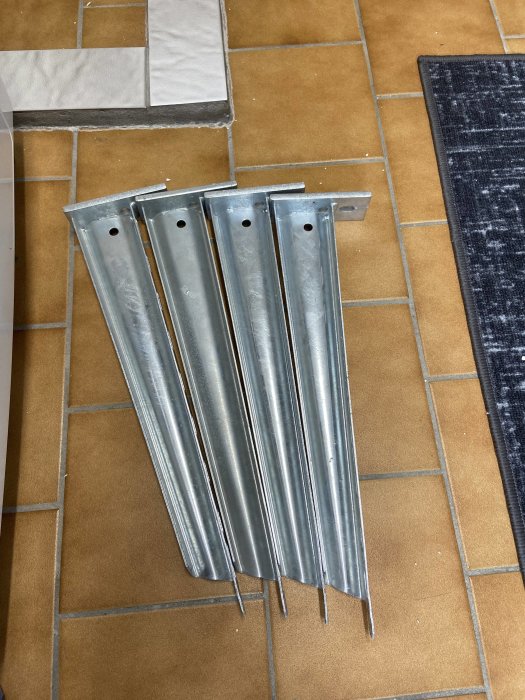 Fyra metallkonsoler ligger på ett terrakottafärgat klinkergolv bredvid en matta, redo för väggmontage.