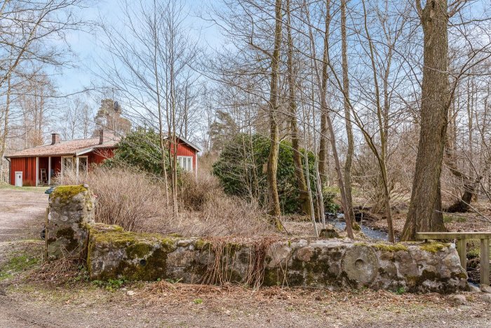 Rött torp omgivet av lövträd med en bäck synlig genom tomten, gamla stenfundament i förgrunden.