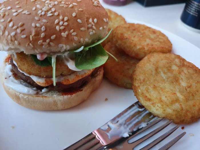 Hamburgare med sesamfrön, sallad och sås, serverad med rösti-tallrikar och en gaffel.