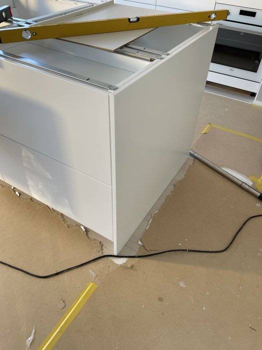 Köksskåpsinstallation med täcksida och vattenpass, omgiven av byggmaterial och verktyg.