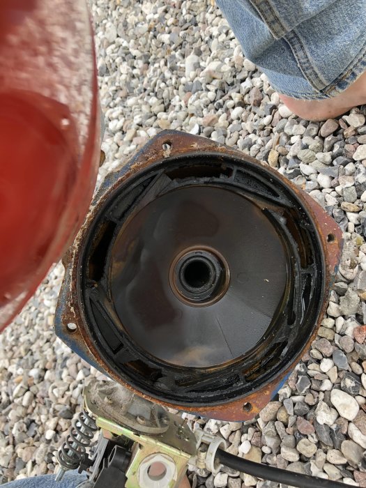 Öppen vattenpump med synligt pumphjul, rost och smuts på ytan, placerad på grusunderlag, nära en persons jeansklädda ben.