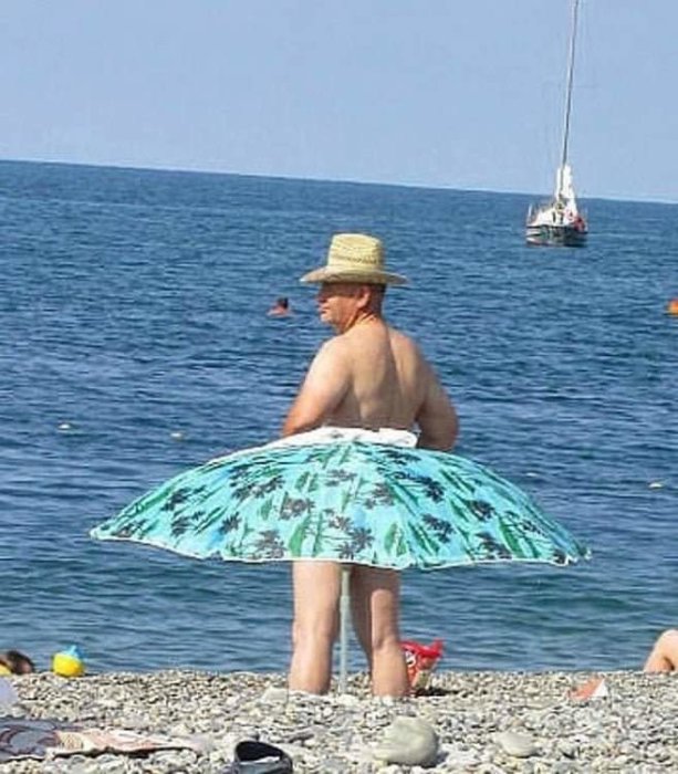 Person med halmhatt och blommig uppblåsbar flytring runt midjan vid strandkanten, med havet och segelbåt i bakgrunden.