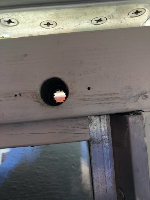 Hål borrat i ett träelement nära en metallkarm med synliga skruvskallar, antyder en dörr- eller fönsterinstallation.