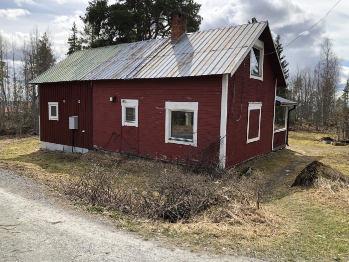 Rött trähus med plåttak och vita fönsterkarmar, omgivet av torrt gräs och träd.