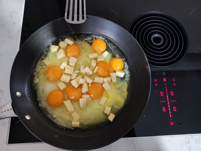 Stekpanna på spis med sex fräsande ägg och kuber av ost, köksredskap, på en elektrisk spis med digital display.