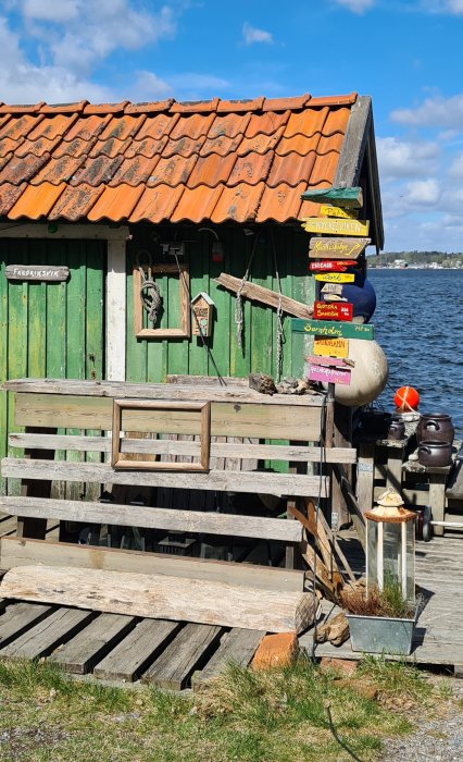Grönmålat sjöbod med rostigt tegeltak och träbänk, vid vattnet, med vägvisarskyltar och maritima dekorationer.