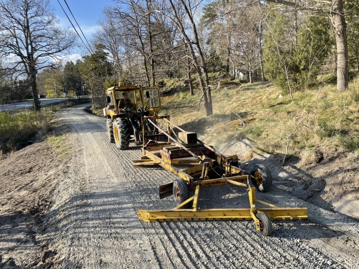 Gul traktor som backar med vägsladd på en grusväg omgiven av träd.