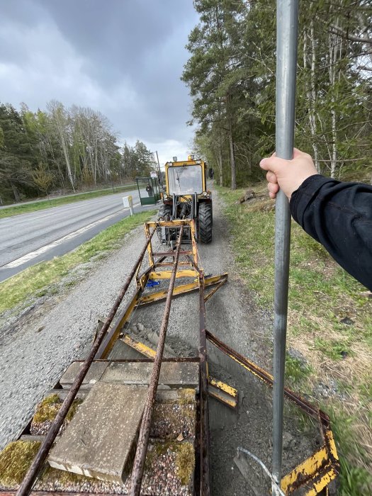 En person som backar med en traktor och tom vagn längs en smal vägkant.