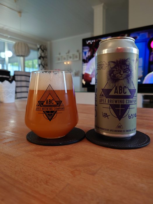 Ölglas med Apex Brewing Company-logotyp fyllt med öl och en ölburk med motiv på en bordsskiva.