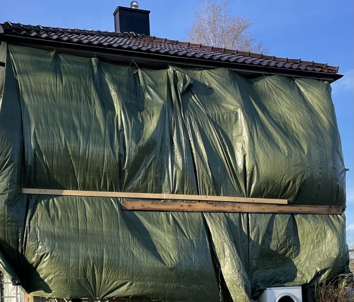 Grön pressening hänger på en träställning för fasadskydd, med släppta öljetter och skador från vind.