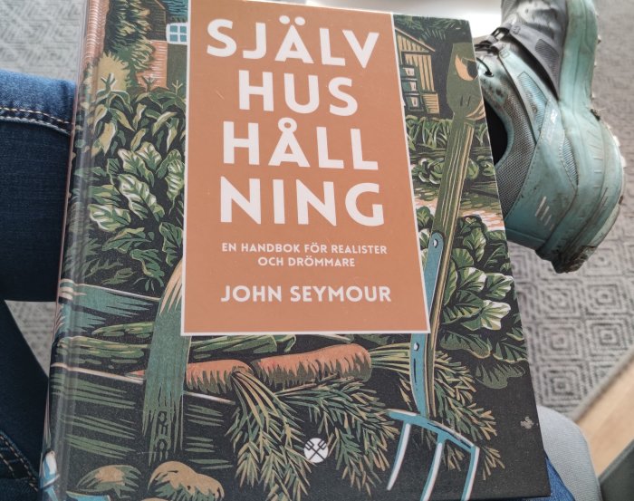 Bok med titeln "SJÄLVHUSHÅLLNING" av John Seymour i någons knä bredvid en sneaker på ett ben i blåjeans.
