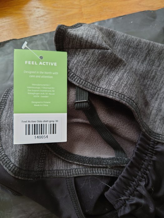 Etikett på en grå 'Feel Active Oslo shirt' från Arken Zoo med storlek och produktkod.