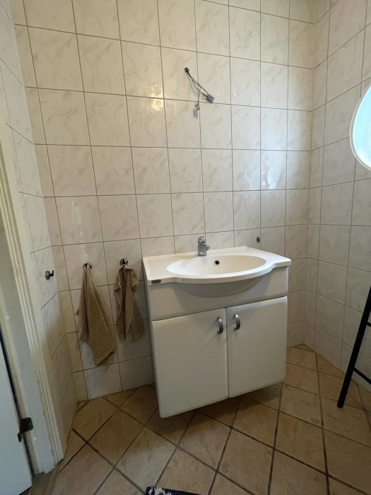 Ett badrum med kaklade väggar och mörkbrunt klinkergolv med en vit badrumskommod och en silverfärgad handdukshängare.