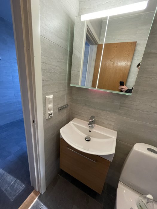 Badrum med träkommod, handfat, toalett och väggmonterad spegel med belysning.