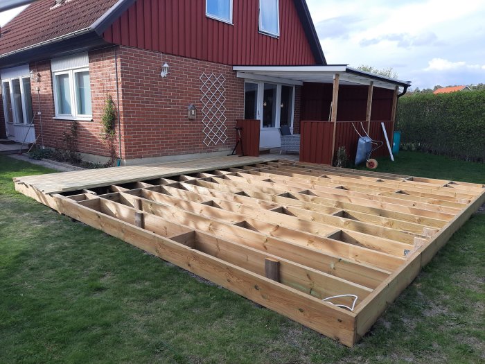Pågående byggnation av träaltan bredvid ett rött tegelhus med öppen trästomme synlig.
