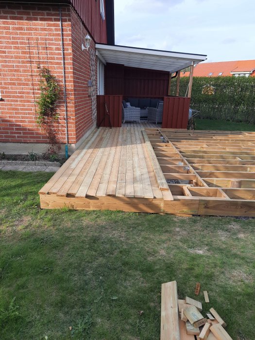 Byggnation av träaltan vid röd tegelvilla med verktyg och material synliga på gräset.