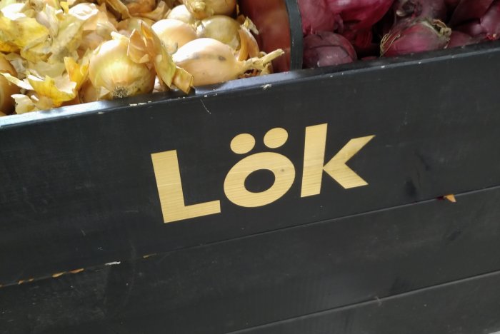 En svart låda med ordet "LÖK" i guldiga bokstäver, fylld med gula och röda lökar.