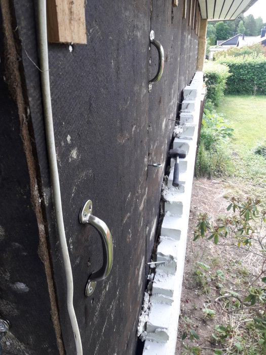 Sidosikt av en husfasad vid en utgrävning för garage med synlig geotextil och påbörjad grundläggning.