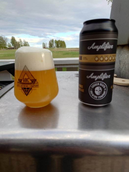 Ölglas fyllt med DIPA och en suddig burk från Sudden Death på ett utomhusbord med landskap i bakgrunden.