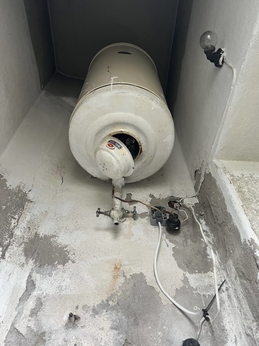 En gammal varmvattenberedare placerad på ett betonggolv med kopplade rör och kranar synlig, ett eluttag och en lampa på väggen.