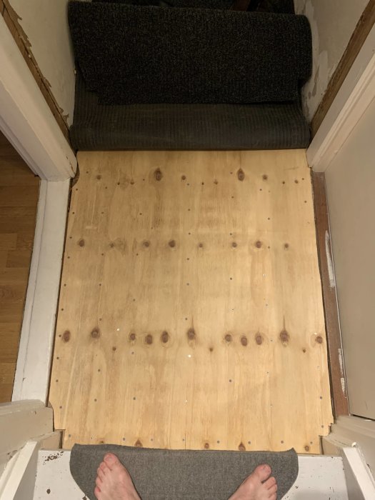Renoverat golv med ny plywood vid en trappa, synliga skruvhuvuden, i förgrunden en persons fötter.