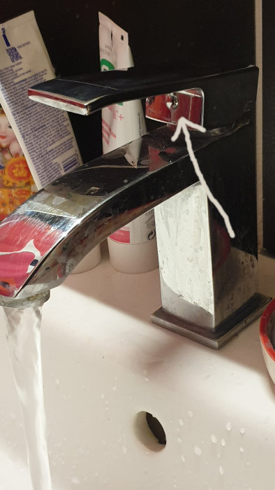 Kromad blandare med löst handtag och vattenflöde, pil pekar på täckplugg, stänk och badrumstillbehör i bakgrunden.