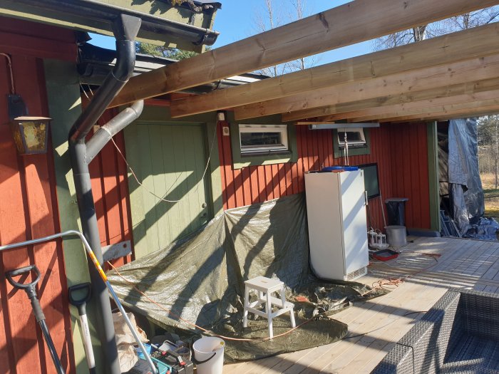 Byggarbetsplats med nyinstallerade reglar på pergola, och befintligt stuprör vid husvägg.