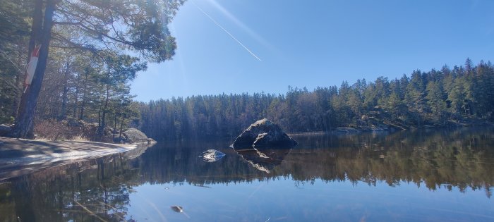 Sjö i Vegalla naturreservat med spegelblank yta, omgiven av barrskog och klippor en solig vårdag.