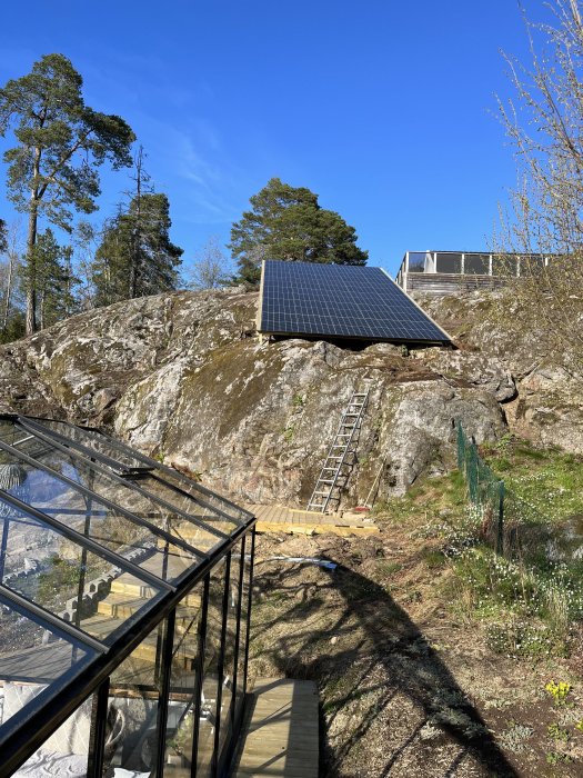 Solpaneler monterade på ett klippigt landskap med träd runtomkring och en stege som leder upp till panelerna.