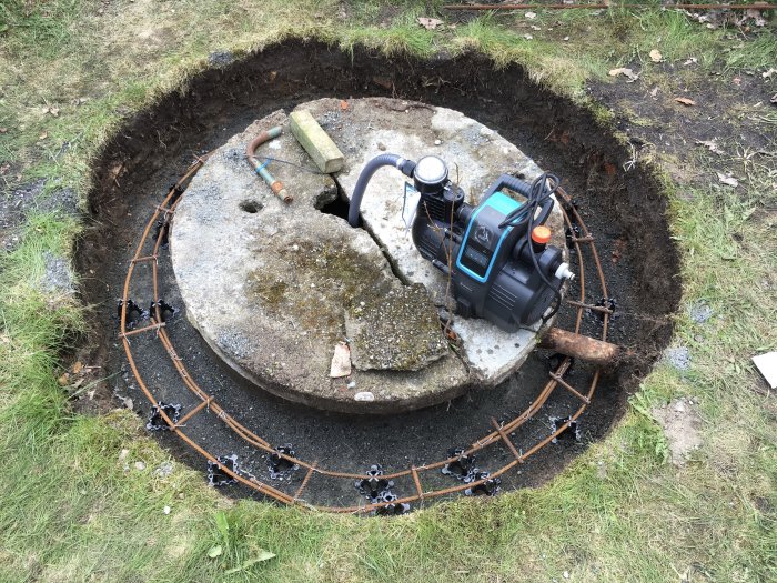 Uppgrävd cirkulär grop runt en betongbrunn med en pump ovanpå, omgiven av armeringsjärn och temporära mätmarkeringar.