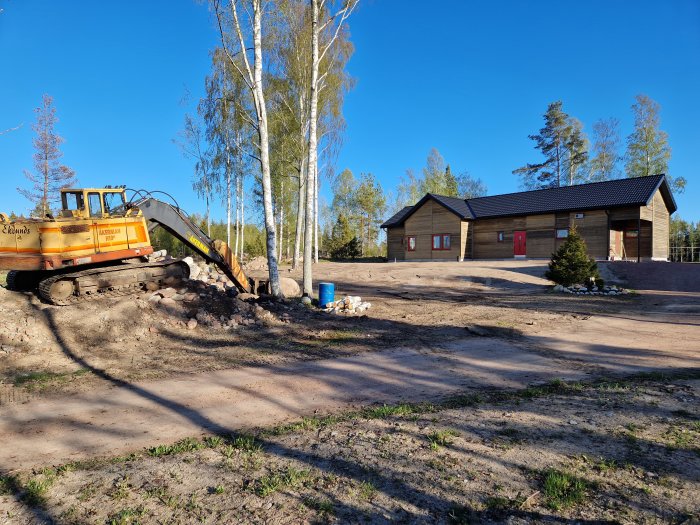 Ett grävfordon framför en nybyggd enplansvilla med planerad mark och björkar under blå himmel.