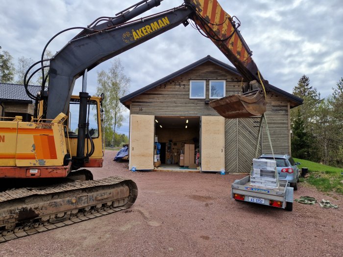 Grävmaskin lyfter byggmaterial på pall i släpvagn framför garage på en uppfart.