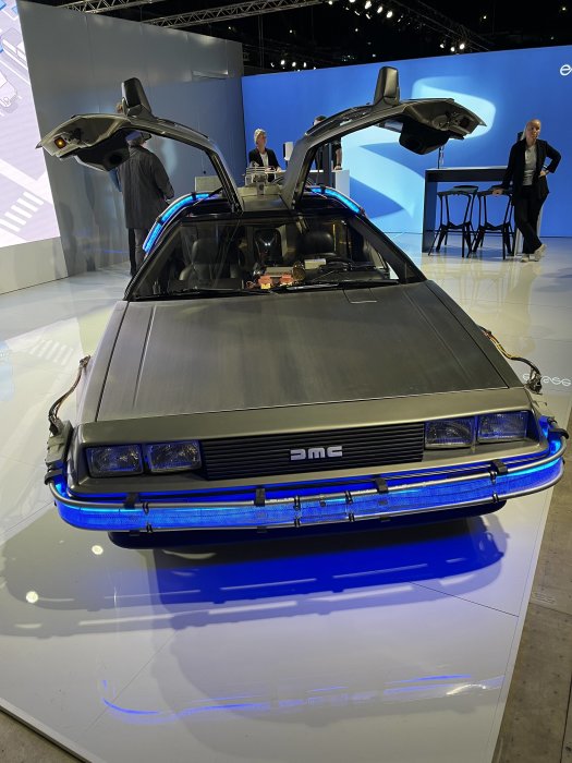 DeLorean DMC-12 med öppna gångjärnsdörrar på elbilsmässa, okänt pris och dragkroksinformation.