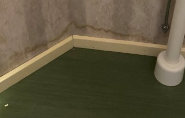 Vägg med fuktskador nära golvet bredvid en vit pelare, vilket tyder på vattenskada.