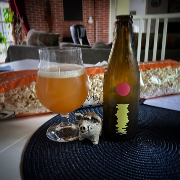 Ölglas på bord med öppnad flaska och en spargris, förgrunden, och popcornpåse och inredning i bakgrunden.