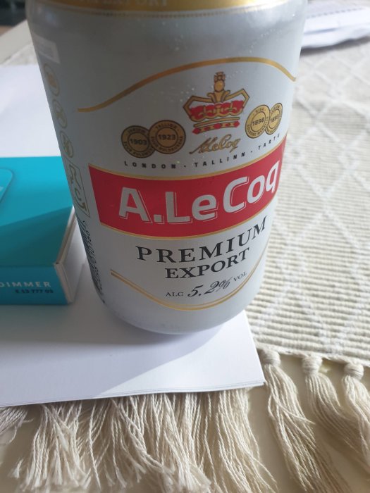 Närbild på en A. Le Coq Premium Export ölburk på ett bord med en blå tändsticksask i bakgrunden.
