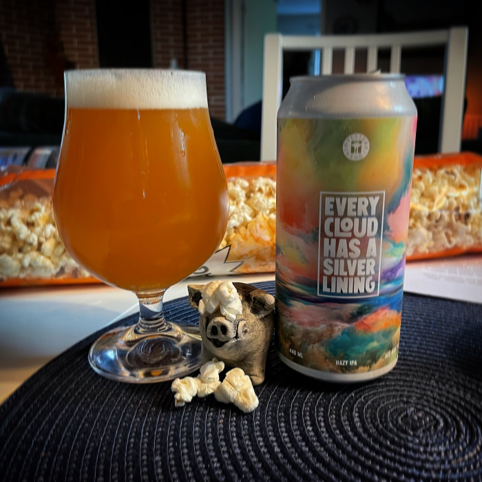 Ölglas med grumligt öl bredvid en burk med texten "Every Cloud Has a Silver Lining" och en liten trägris på ett bord.