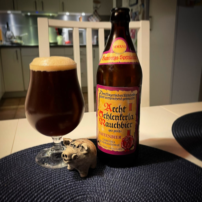 En ölflaska bredvid ett ölglas på ett bord med en liten trägrisfigur i förgrunden.