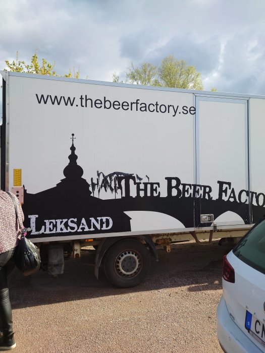 Lastbil med reklam för "The Beer Factory" och webbadress, parkerad bredvid personbil.