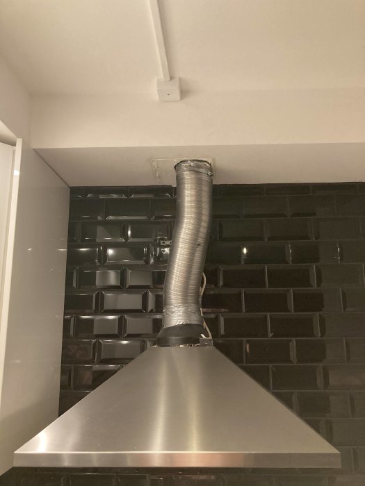 Köksfläkt ansluten till flexibel ventilationsslang mot avluftskanal i taket över mörka kakelplattor.