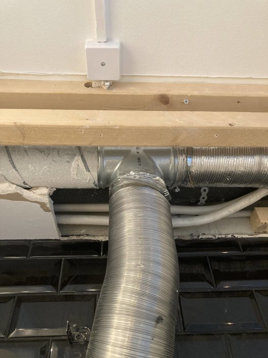 Flexibel ventilationskanal ansluten till en T-koppling under ett köksskåp, bredvid en elledning och träreglar.