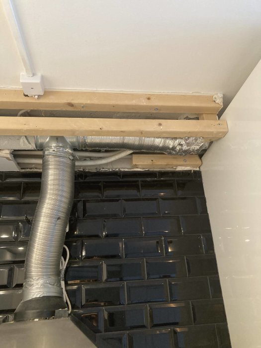 Kök med öppen ventilationsschakt, T-koppling, flexibel slang och svarta kakelväggar.