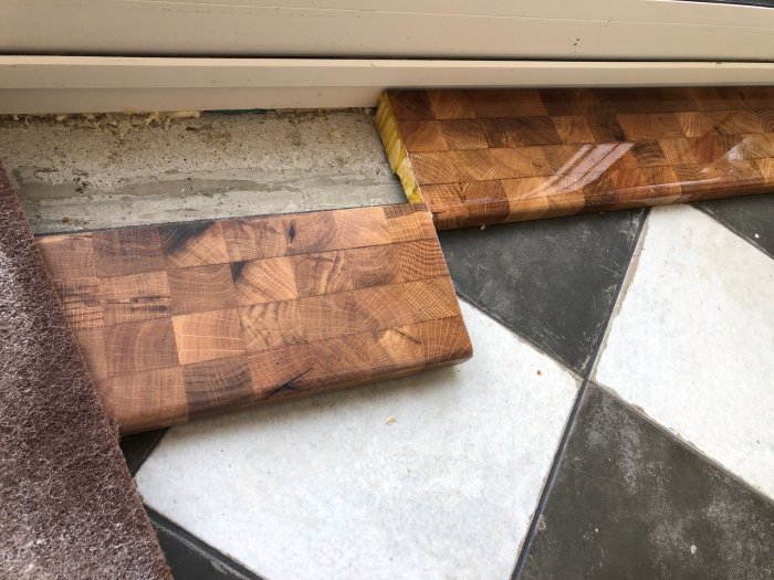 Handgjord trätröskel med kubbmönster vid en dörröppning, mellan matta och klinkergolv.