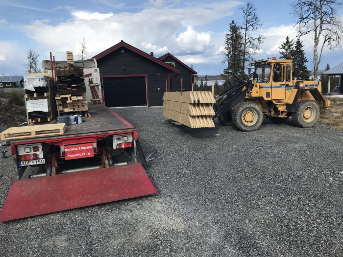 Lastbil fullastad med byggmaterial och hjullastare bredvid på grusplan.
