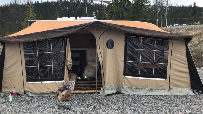 Förtält på husvagn uppställt på grus, omgivet av skog och med byggmaterial synligt inuti.