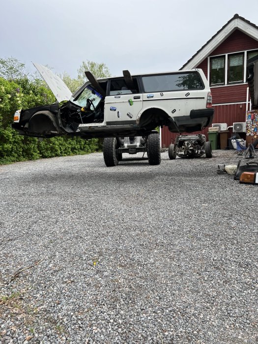 Upphöjd pickup med öppen huva på grus framför röd stuga, diverse saker runtomkring.