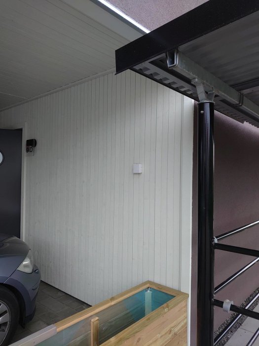 Del av carport med vit panelvägg, takkonstruktion och en del av en bil.